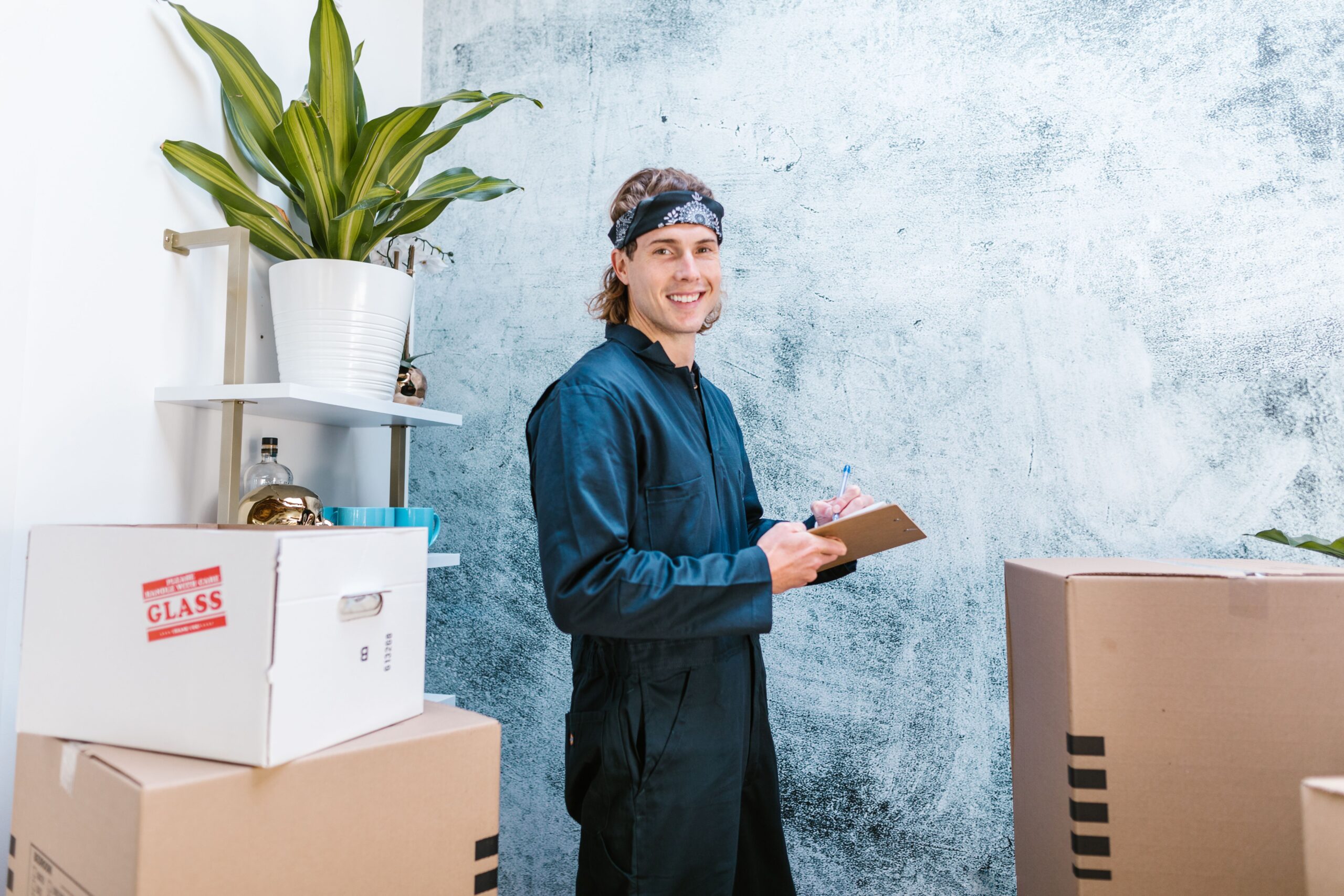 A smiling worker in coveralls checks cardboard boxes indoors, with plants in the background.